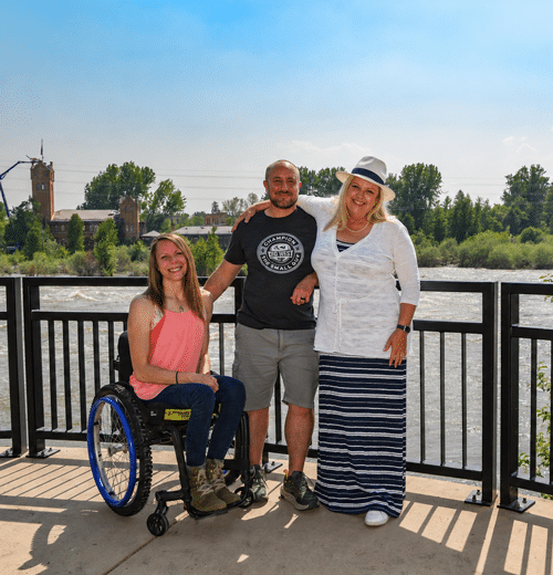 photo of three people. Woman in a white shirt and blue and white striped skirt. She's also wearing a sun hat. There's a man in the middle wearing a grey t-shirt and tan cargo shorts. There's a woman in a wheelchair wearing a pink tank top and jeans. 
